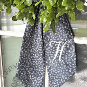 May include: A decorative wreath with lush green leaves and a navy blue fabric bow. The bow features a white floral pattern and a large white letter "H". The text "CEDAR THREAD CO" is visible on the bottom left.