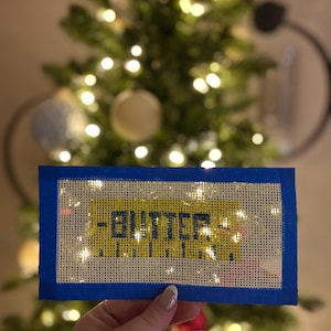 May include: A rectangular needlepoint with the word "-BUTTER-" in black letters on a yellow background, framed by a blue border. The needlepoint is illuminated by small, twinkling lights. The background is a blurred Christmas tree.