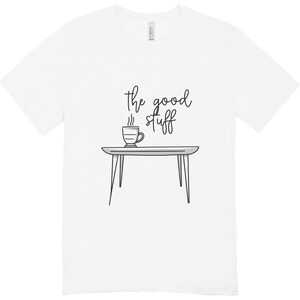 Könnte beinhalten: Weißes T-Shirt mit einer skurrilen Illustration. Das Design zeigt einen kleinen Tisch mit einer dampfenden Kaffeetasse und dem handgeschriebenen Text "the good stuff". Das Shirt ist aus einem weichen Material.