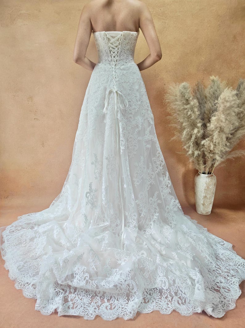 Puede incluir: Vestido de novia blanco sin tirantes de encaje con una larga cola. La parte trasera del vestido presenta un cierre tipo cors&eacute; con un lazo. El vestido est&aacute; hecho de encaje delicado y es largo.