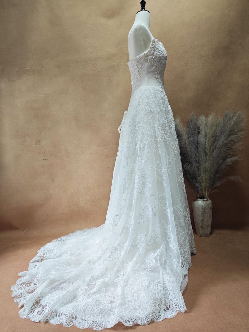 Puede incluir: Un vestido de novia blanco sin tirantes con intrincados detalles de encaje. El vestido presenta un corpi&ntilde;o ajustado y una falda fluida que se extiende en una larga cola. El vestido se exhibe en un maniqu&iacute;.