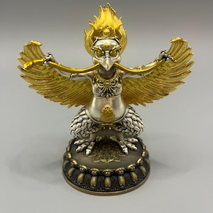 Può includere: Una statuetta metallica di una creatura mitica simile a un uccello con corpo argentato e ali, cresta e serpente dorati tenuti tra gli artigli. La figura poggia su una base circolare color bronzo scuro con un intricato motivo a petali di loto.