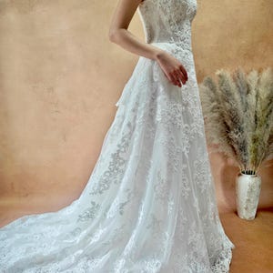 Puede incluir: Vestido de novia blanco, sin tirantes, con intrincados detalles de encaje. El vestido presenta una falda fluida y una larga cola. El vestido se muestra sobre un fondo neutro, resaltando los delicados patrones de encaje y la elegancia general.