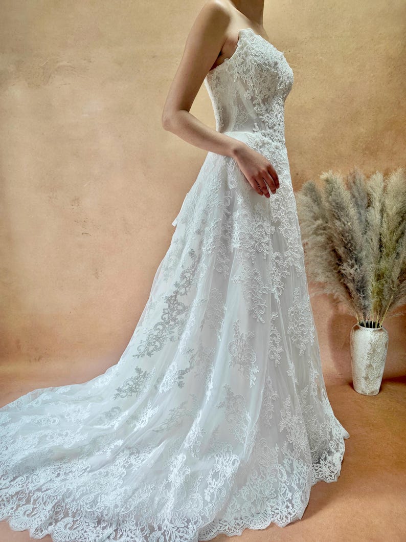 Puede incluir: Vestido de novia blanco, sin tirantes, con intrincados detalles de encaje. El vestido presenta una falda fluida y una larga cola. El vestido se muestra sobre un fondo neutro, resaltando los delicados patrones de encaje y la elegancia general.