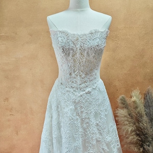 Puede incluir: Vestido de novia blanco sin tirantes de encaje, exhibido en un maniqu&iacute;. El vestido presenta intrincados detalles de encaje floral en el corpi&ntilde;o y la falda. El vestido tiene una silueta fluida en forma de A.