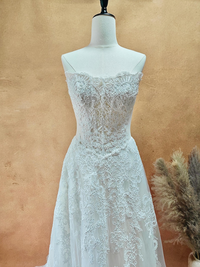 Puede incluir: Vestido de novia blanco sin tirantes de encaje, exhibido en un maniqu&iacute;. El vestido presenta intrincados detalles de encaje floral en el corpi&ntilde;o y la falda. El vestido tiene una silueta fluida en forma de A.