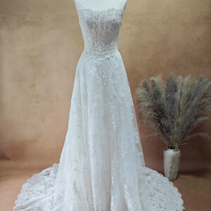 Puede incluir: Vestido de novia blanco con encaje. El vestido presenta un corpi&ntilde;o ajustado, tirantes finos y una falda fluida que se extiende en una cola. El vestido se exhibe en un maniqu&iacute;.