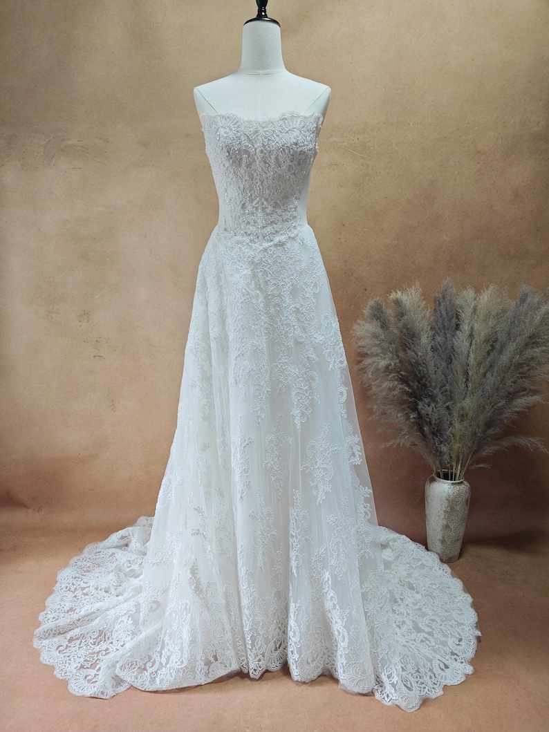 Puede incluir: Vestido de novia blanco con encaje. El vestido presenta un corpi&ntilde;o ajustado, tirantes finos y una falda fluida que se extiende en una cola. El vestido se exhibe en un maniqu&iacute;.