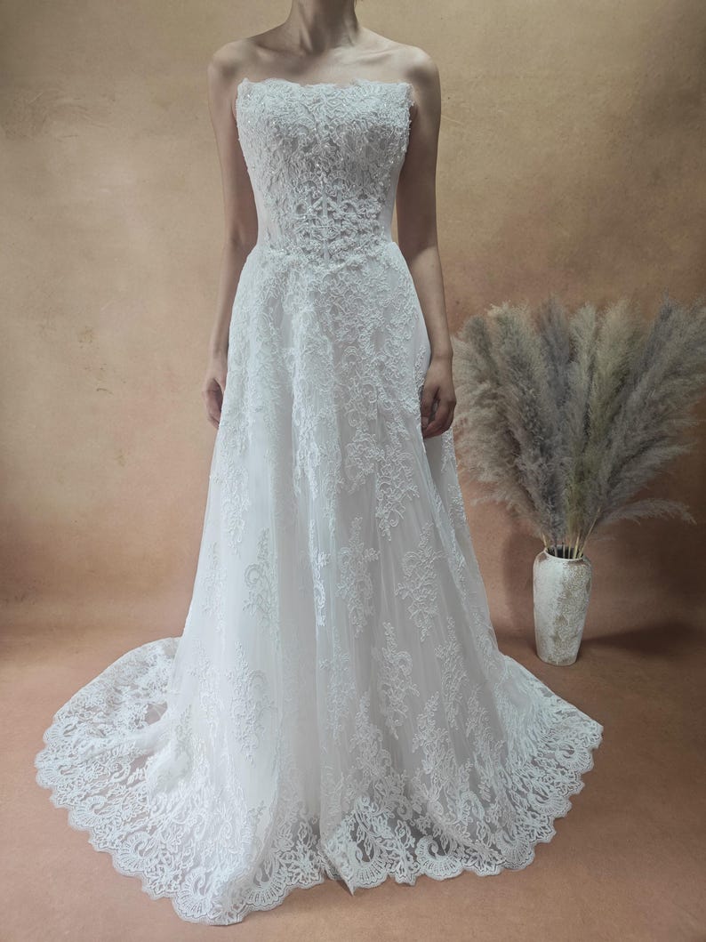 Puede incluir: Vestido de novia blanco sin tirantes con encaje. El vestido tiene una falda fluida y una cola. El corpi&ntilde;o est&aacute; adornado con intrincados patrones de encaje floral. Vestido largo, ideal para una boda o evento formal.