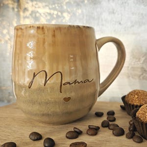 Könnte beinhalten: Eine beige Keramiktasse mit dem Wort "Mama" in Schreibschrift und einem Herzsymbol. Die Tasse steht auf einer Holzoberfläche mit Kaffeebohnen und zwei Schokoladentrüffeln in Papierförmchen.