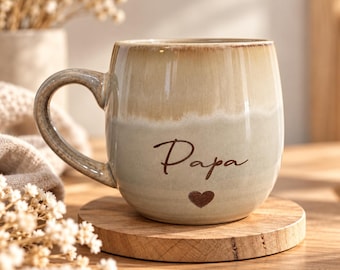 Könnte beinhalten: Ein Keramikbecher mit zweifarbiger Glasur, bestehend aus einem beigen oberen Teil und einem hellgrauen unteren Teil. Das Wort Papa ist in dunkelbrauner Schreibschrift über einem kleinen dunkelbraunen Herzsymbol auf der Vorderseite aufgedruckt.