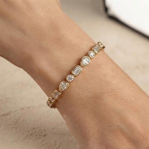 May include: A gold bracelet featuring a series of alternating clear gemstones in various cuts, including round, pear, and emerald shapes. Each stone is set in a polished gold bezel setting, creating a continuous line of sparkling stones around the wrist.