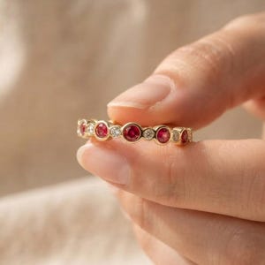 Op de afbeelding: Een gouden ring met een afwisselend patroon van ronde, in een zetting geplaatste rode robijnen en heldere witte diamanten. De edelstenen zijn in een doorlopende rij langs de metalen band geplaatst, wat zorgt voor een cirkelvormig ontwerp.