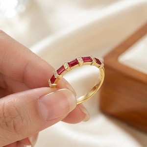 May include: A gold band ring featuring a series of alternating rectangular red rubies and small round clear diamonds set in a row along the top half of the band. The metal has a polished yellow finish, and the stones are held in place by metal prongs.