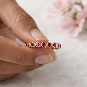 Puede incluir: Una alianza de oro con una hilera continua de rubíes rojos redondos engastados en bisel. El anillo está construido con un marco de metal dorado pulido que asegura cada piedra preciosa individual en un engaste circular, creando un diseño uniforme.