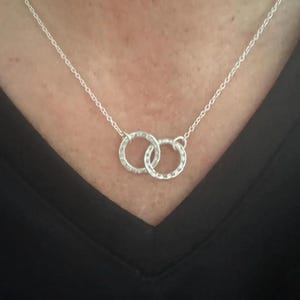 Puede incluir: Un collar plateado con dos anillos texturizados e entrelazados. Los anillos están conectados y suspendidos de una fina cadena. El collar se muestra sobre un fondo negro.