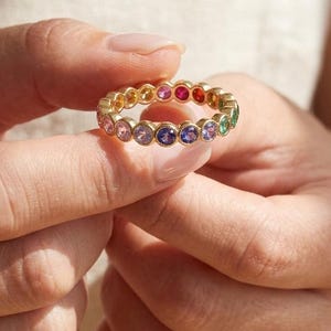 May include: A gold eternity band featuring a continuous row of bezel-set round gemstones in a vibrant rainbow spectrum, including shades of pink, red, orange, yellow, green, blue, and purple, creating a colourful circular design.