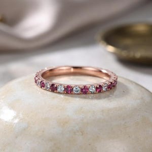 Könnte beinhalten: Ein Ewigkeitsring aus Roségold mit einem abwechselnden Muster aus rund geschliffenen, tiefroten Rubinen und klaren, farblosen Diamanten, die in einer durchgehenden Reihe um den gesamten Umfang des dünnen, polierten Metallrings gefasst sind.