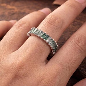 May include: A silver-toned band ring featuring a continuous row of rectangular, baguette-cut green moss agate stones. The stones are set side-by-side in a prong setting, creating a full eternity style band with visible natural inclusions.