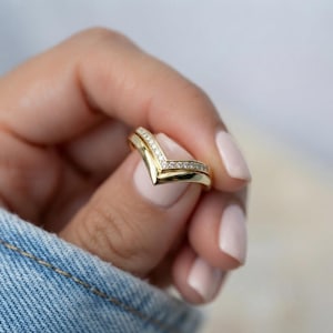 以下が含まれることがあります： 上端に沿って小さな透明な輝く石が一列に並んだ、ゴールドのV字型シェブロンリング。バンドは磨き上げられた滑らかな仕上げで、指の上で独特の角度のある形状を作り出す尖ったデザインが特徴です。