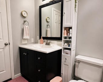 May include: A modern bathroom with a black vanity, white countertop, and a large black-framed mirror. A tall white cabinet provides storage, and a pink rug and trash can add a pop of color. A gold clock and towel rack are mounted on the wall.