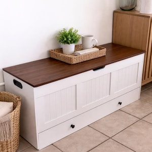 May include: A white storage bench with a dark brown wooden top. The bench has two drawers with black knobs and a slatted design on the front. A woven tray with a plant, mug, and book sits on top.