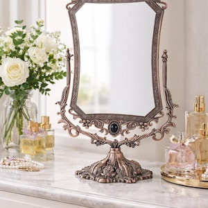 May include: Ornate bronze-colored vanity mirror with a decorative floral base and frame. The mirror has an octagonal shape and is embellished with dark oval accents. The mirror is on a white marble surface with perfume bottles and a bouquet of white roses.