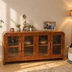 May include: A wooden sideboard with four glass-fronted doors, showcasing interior shelves with dishes. The cabinet has a warm, natural wood finish and gold-toned handles. A framed photo and decorative items sit on top.