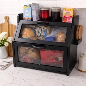 May include: A black wooden bread box with two compartments and glass doors. The top shelf holds jars, juice boxes, and a box of raspberry-flavored vitamin C. The bread box is on a white countertop.