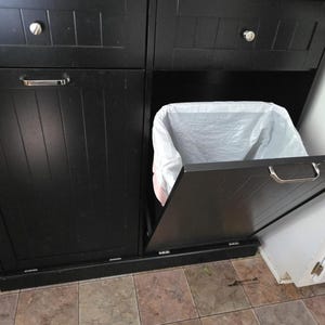 May include: Black cabinet with two doors and a pull-out trash bin. The cabinet doors have silver handles and knobs. A white trash bag is visible inside the open bin. The cabinet is on a tiled floor.