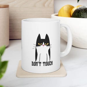 Op de afbeelding: Witte keramische mok met een zwart-witte kattenillustratie en gele ogen. De mok heeft de tekst "DON'T TOUCH" in zwarte letters. De mok staat op een beige onderzetter.