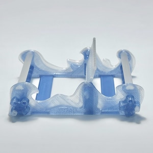 Puede incluir: Un soporte de exhibición azul translúcido impreso en 3D con dos paneles laterales en forma de onda conectados por dos varillas horizontales. La estructura presenta una textura en capas y un divisor central vertical para sostener objetos.