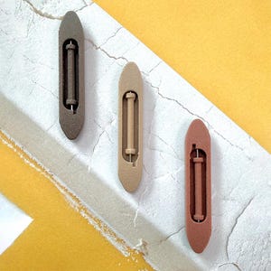 Peut inclure: Trois navettes de tissage en bois, dans des tons de marron, beige et brun rougeâtre, sont disposées sur une surface texturée blanche et jaune. Chaque navette présente une ouverture centrale avec un composant cylindrique.
