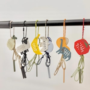 Puede incluir: Una colección de llaveros coloridos con varias formas, incluyendo una oveja, un plátano y un dinosaurio. Cada llavero está unido a una cadena de cuentas y un cordón anudado. Los llaveros son en tonos de amarillo, rojo, azul y verde.