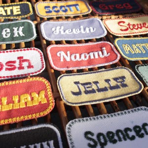 May include: A collection of embroidered name patches in various colors, including red, blue, yellow, and green. Each patch features a different name in a contrasting color, such as "Kate," "Josh," and "William." The patches are arranged on a wooden surface.