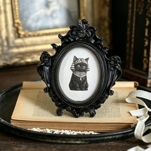 May include: A black oval picture frame with ornate detailing holds a black and white illustration of a cat wearing an Egyptian headdress. The frame sits on a vintage book and a black tray.
