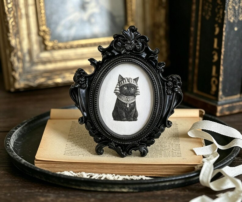 May include: A black oval picture frame with ornate detailing holds a black and white illustration of a cat wearing an Egyptian headdress. The frame sits on a vintage book and a black tray.