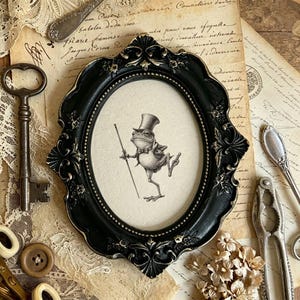 May include: An oval black picture frame with ornate metallic detailing surrounding a central illustration of a frog wearing a top hat and bow tie while holding a walking stick. The frame features intricate floral carvings and a beaded inner edge.