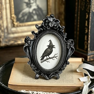 May include: Black oval picture frame with a crow wearing a witch hat perched on a branch. The frame is ornate and sits on a stack of old books. The crow illustration is in black and white.