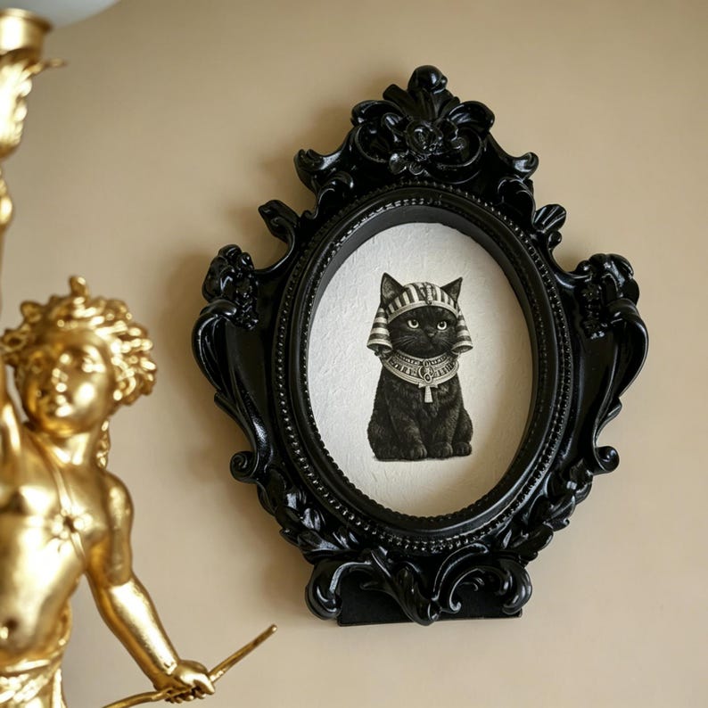 May include: Black oval picture frame with ornate detailing. The frame holds an image of a black cat wearing an Egyptian headdress and collar. The frame is mounted on a beige wall, with a gold statue visible on the left.