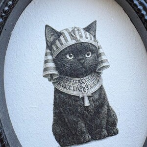 May include: A black cat illustration wearing an Egyptian pharaoh headdress and collar, set within a black oval frame. The artwork is detailed with fine lines, creating a textured appearance. The cat's expression is gentle, and the overall style is reminiscent of vintage art.