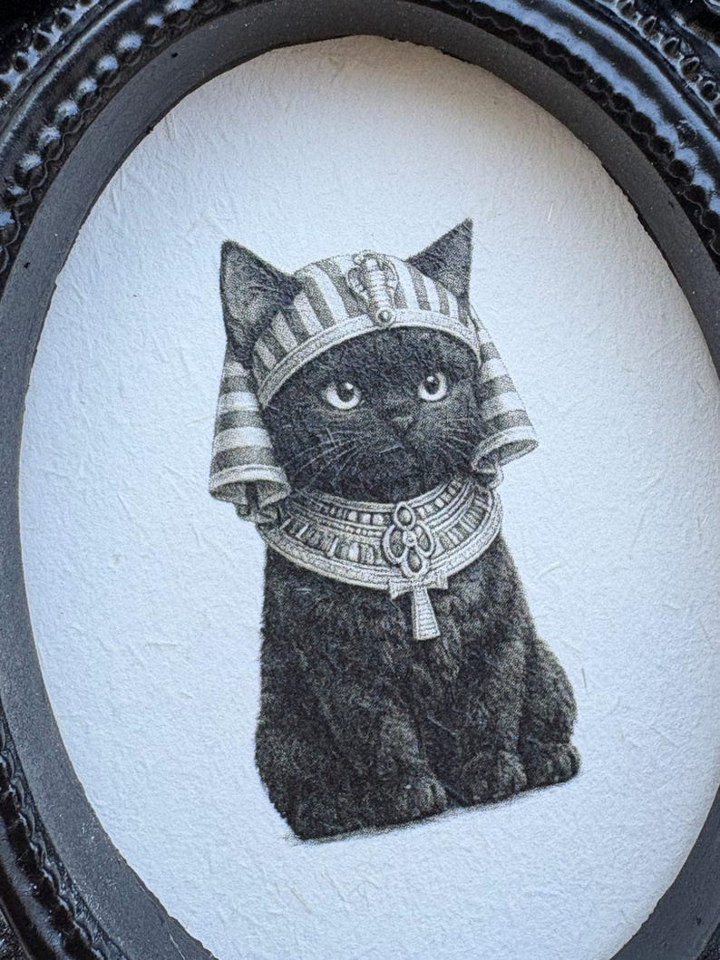 May include: A black cat illustration wearing an Egyptian pharaoh headdress and collar, set within a black oval frame. The artwork is detailed with fine lines, creating a textured appearance. The cat's expression is gentle, and the overall style is reminiscent of vintage art.