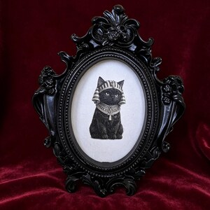 May include: Black framed oval print featuring a black cat in an Egyptian pharaoh headdress and collar. The ornate black frame has decorative flourishes and is set against a deep red velvet background.