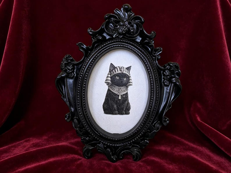 May include: Black framed oval print featuring a black cat in an Egyptian pharaoh headdress and collar. The ornate black frame has decorative flourishes and is set against a deep red velvet background.