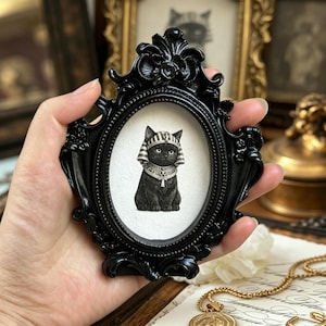 May include: A black framed oval portrait featuring a black cat wearing an Egyptian headdress and collar. The frame is ornate and black. The cat is centered on a white background. The portrait is held in a hand.