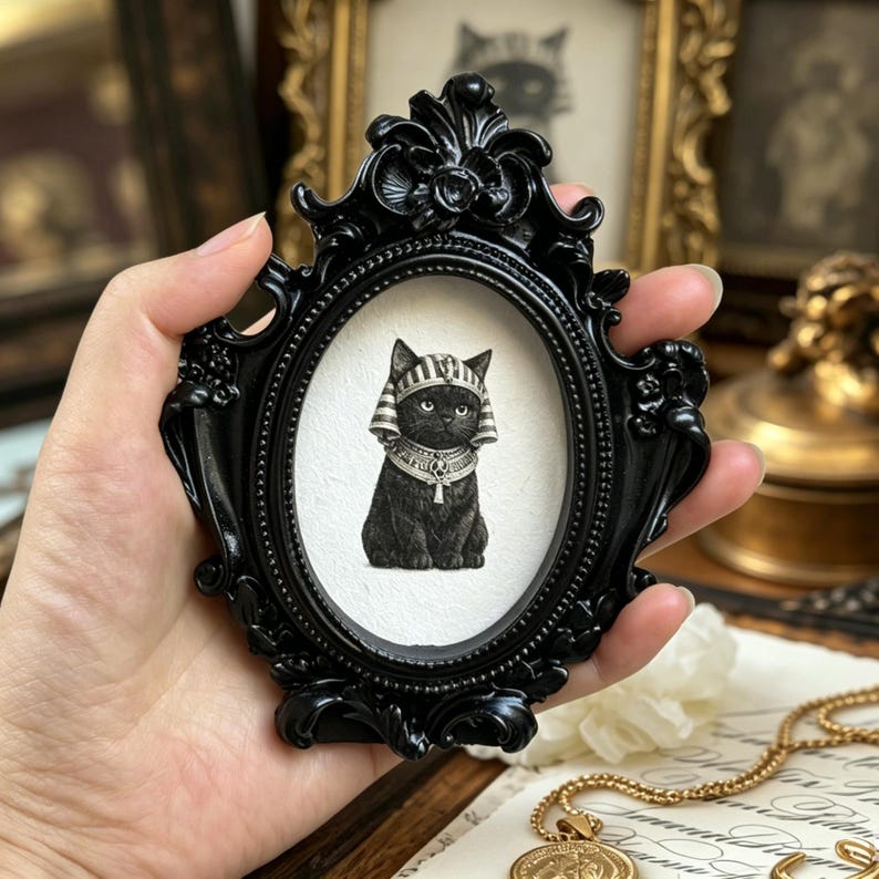 May include: A black framed oval portrait featuring a black cat wearing an Egyptian headdress and collar. The frame is ornate and black. The cat is centered on a white background. The portrait is held in a hand.
