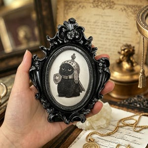Könnte beinhalten: Ein ovaler Bilderrahmen in Schwarz mit einem Bild einer schwarzen Katze in Ritterrüstung. Die Illustration zeigt die Katze mit Schild und Helm. Der Rahmen hat eine dunkle Oberfläche. Der Hintergrund ist neutral.