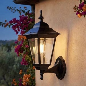 Könnte beinhalten: Eine schwarze Außenwandleuchte aus Metall mit klaren Glasscheiben und einer spitzen Zierkappe. Die Leuchte ist an einer beigen Stuckwand montiert und enthält eine leuchtende Edison-Glühbirne. Ein kleines Schild trägt die Aufschrift Artisan Collection.
