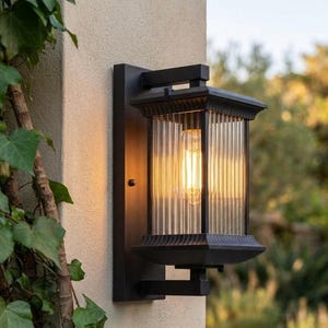 Könnte beinhalten: Eine schwarze Außenwandleuchte mit rechteckigem Rahmen und vertikal gerippten Glasscheiben. Die Leuchte ist an einer hellen Wand montiert und enthält eine einzelne leuchtende Glühbirne, die durch die strukturierten Glasscheiben sichtbar ist.