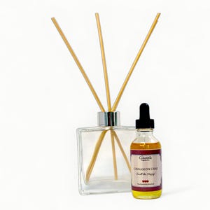 Könnte beinhalten: Ein klarer Glasdiffusor mit Holzstäbchen und eine kleine Glasflasche mit Zimt-Chai-Duftöl. Die Ölflasche hat eine schwarze Pipette und ein Etikett mit der Aufschrift "Cinnamon Chai Smell the Happy!"
